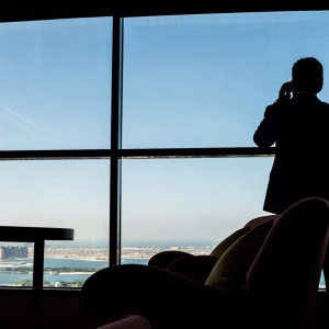 Silhouette of a businessman talking on a phone by a large office window overlooking a coastal city skyline with lounge chairs in the foreground