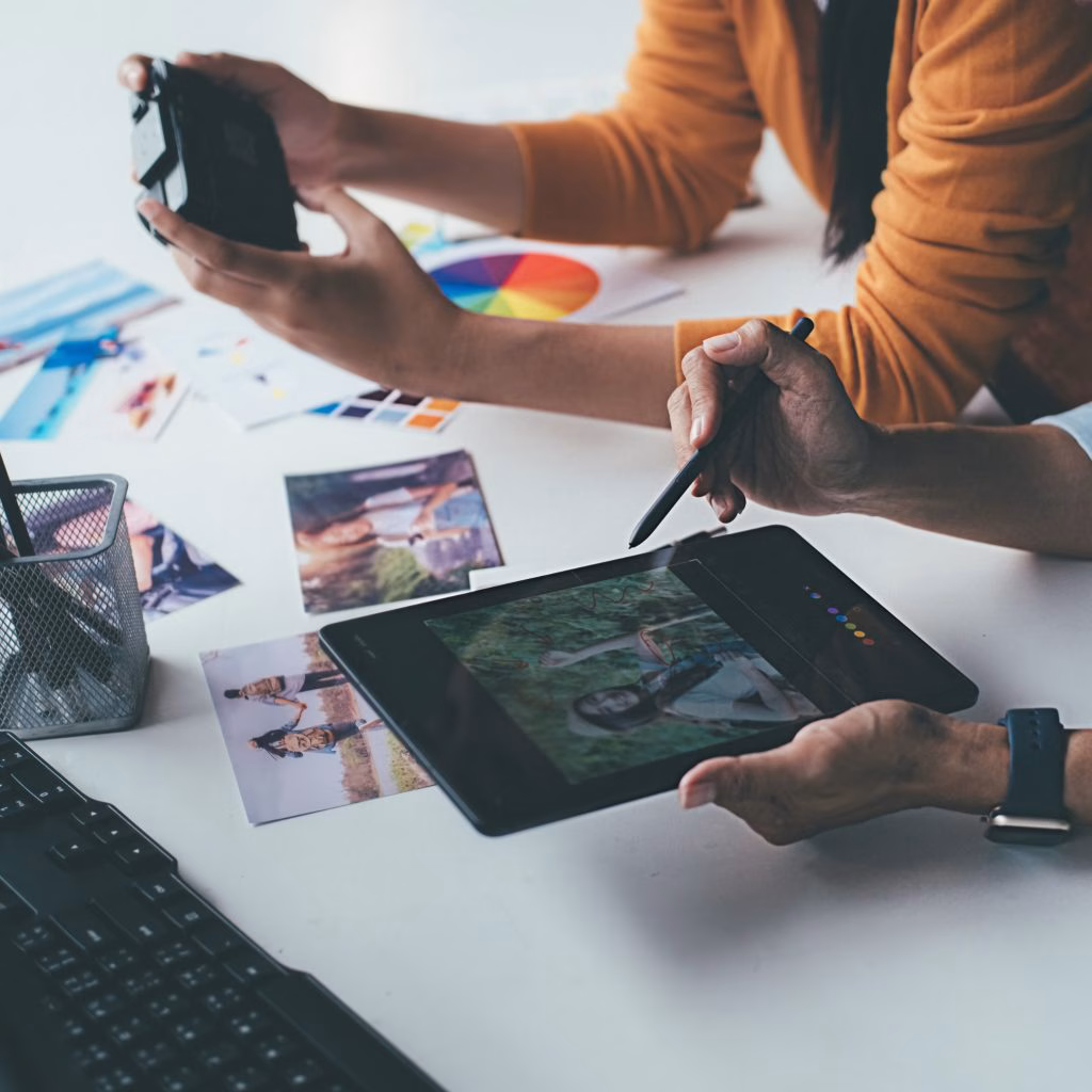 communication Gros plan sur un graphiste utilisant un stylet sur une tablette graphique pour retoucher un portrait, aux côtés d'un collègue tenant un appareil photo, au-dessus d'un bureau où sont disposés des cercles chromatiques et des photos imprimés.