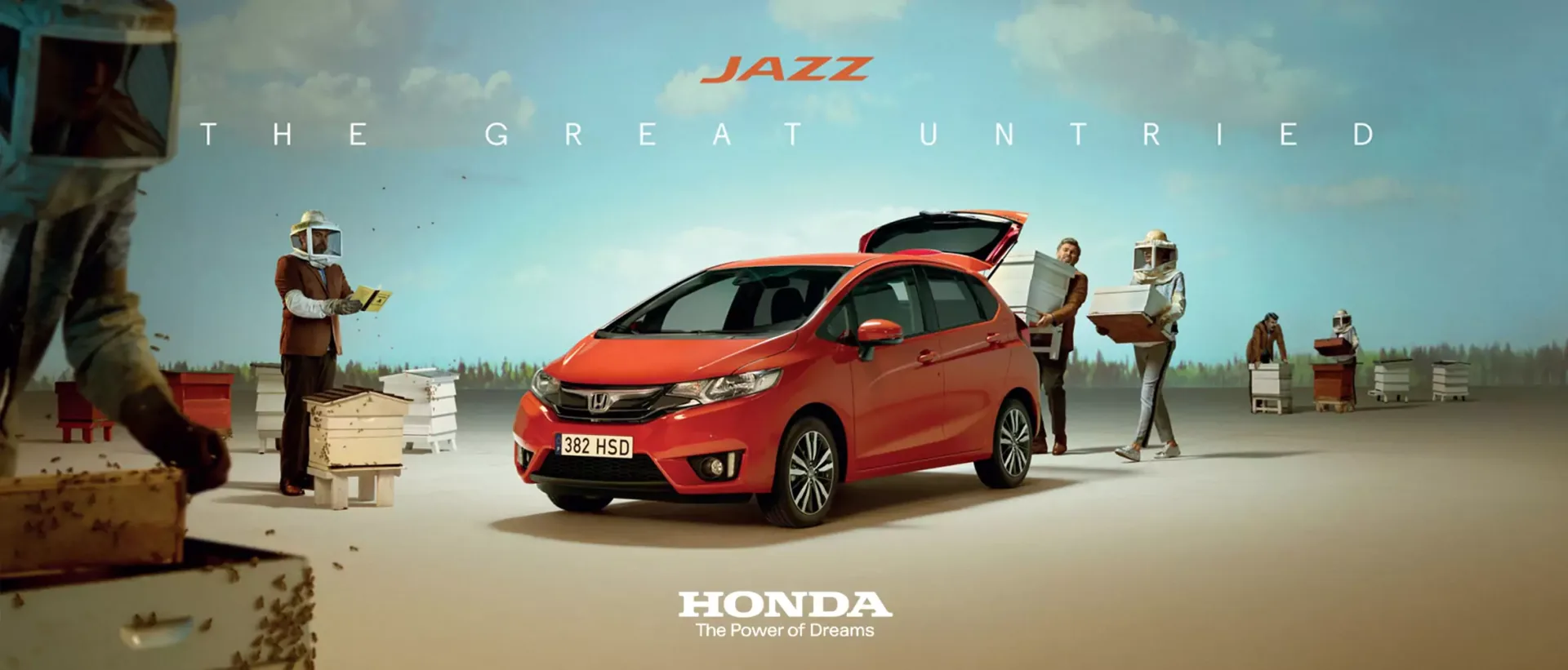 Red Honda Jazz hatchback car parked outdoors surrounded by beekeepers tending to wooden beehives