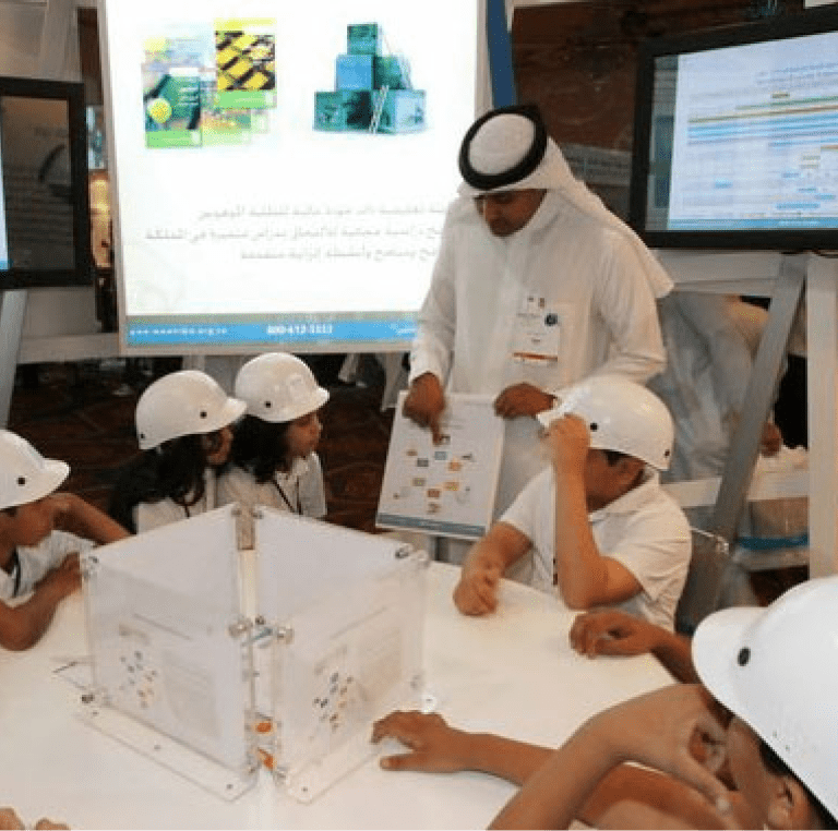 A presenter in traditional Middle Eastern attire pointing to a document during a workshop session with children wearing white safety helmets around a table