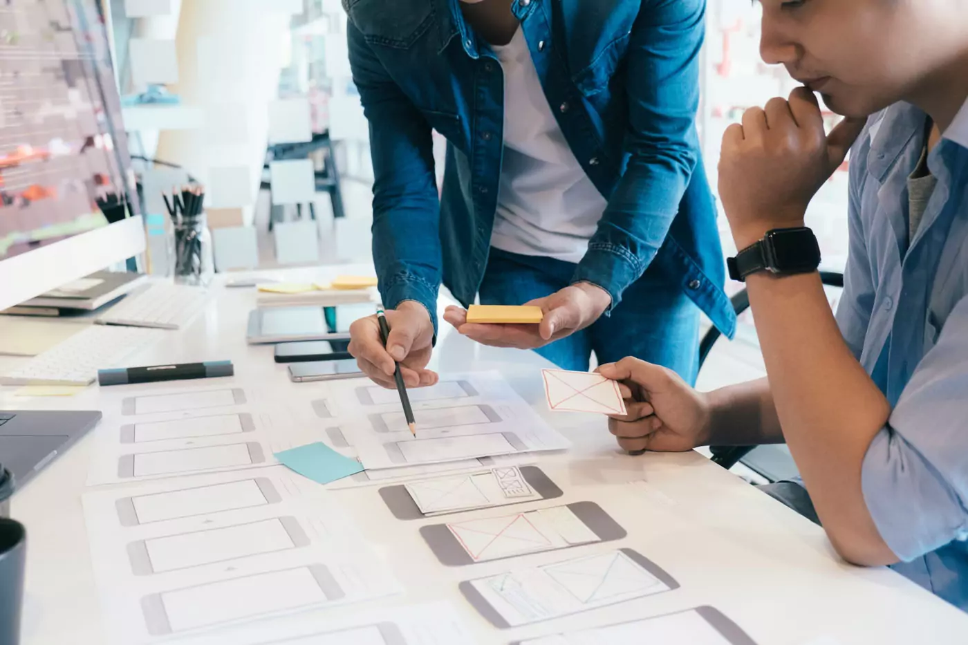 Two designers working together at a desk reviewing printed mobile application wireframe sketches and using sticky notes